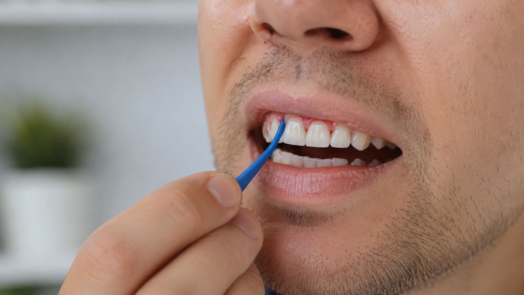 A close-up view of a man using a small blue dental tool to clean between his teeth.