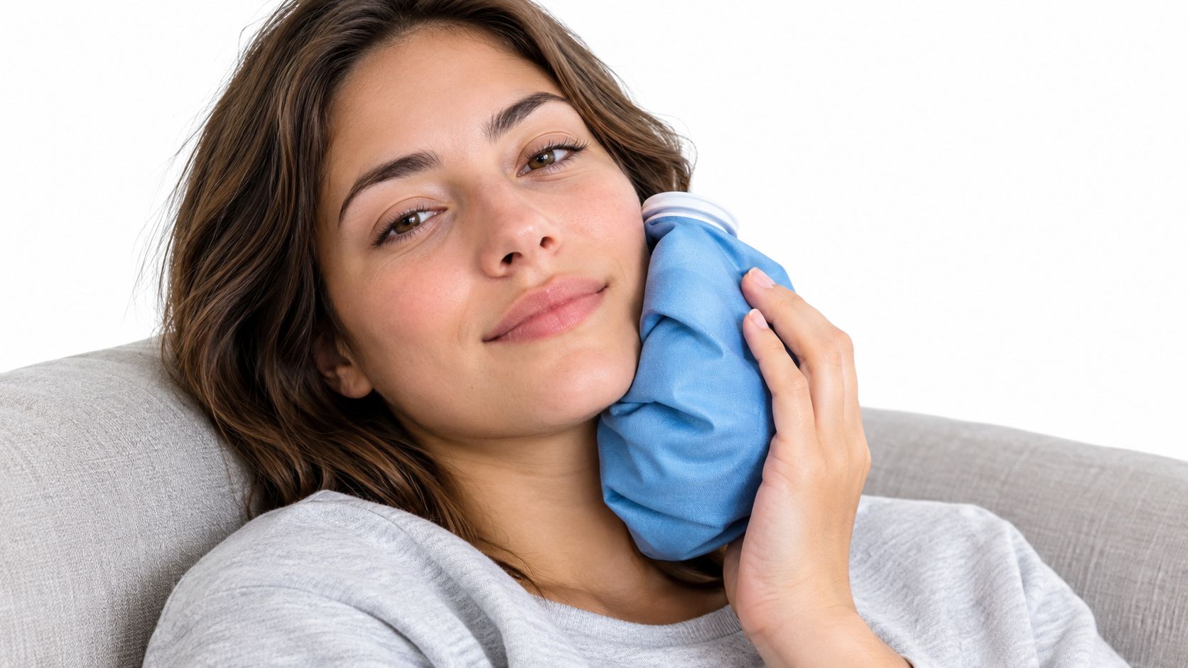 A young woman smiling while resting comfortably with a blue ice pack held against her swollen cheek.